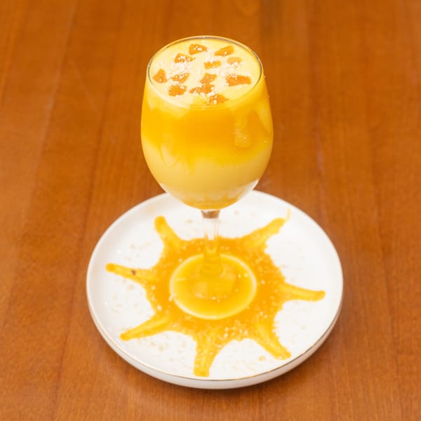 A mango lassi in a wine glass, garnished with mango pieces and sugar, on a white plate.