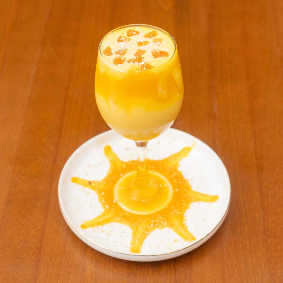 A mango lassi in a wine glass, garnished with mango pieces and sugar, on a white plate.