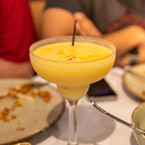 A yellow mango lassi in a margarita glass with a straw, on a table with empty plates.