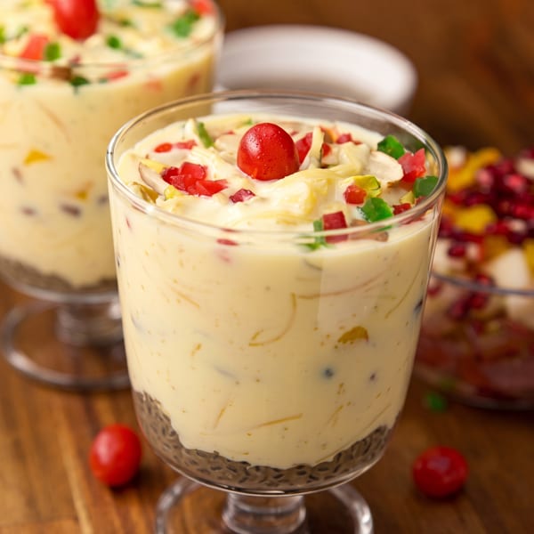 Two glasses of creamy dessert with vermicelli, topped with candied fruits and a cherry on a wooden table.